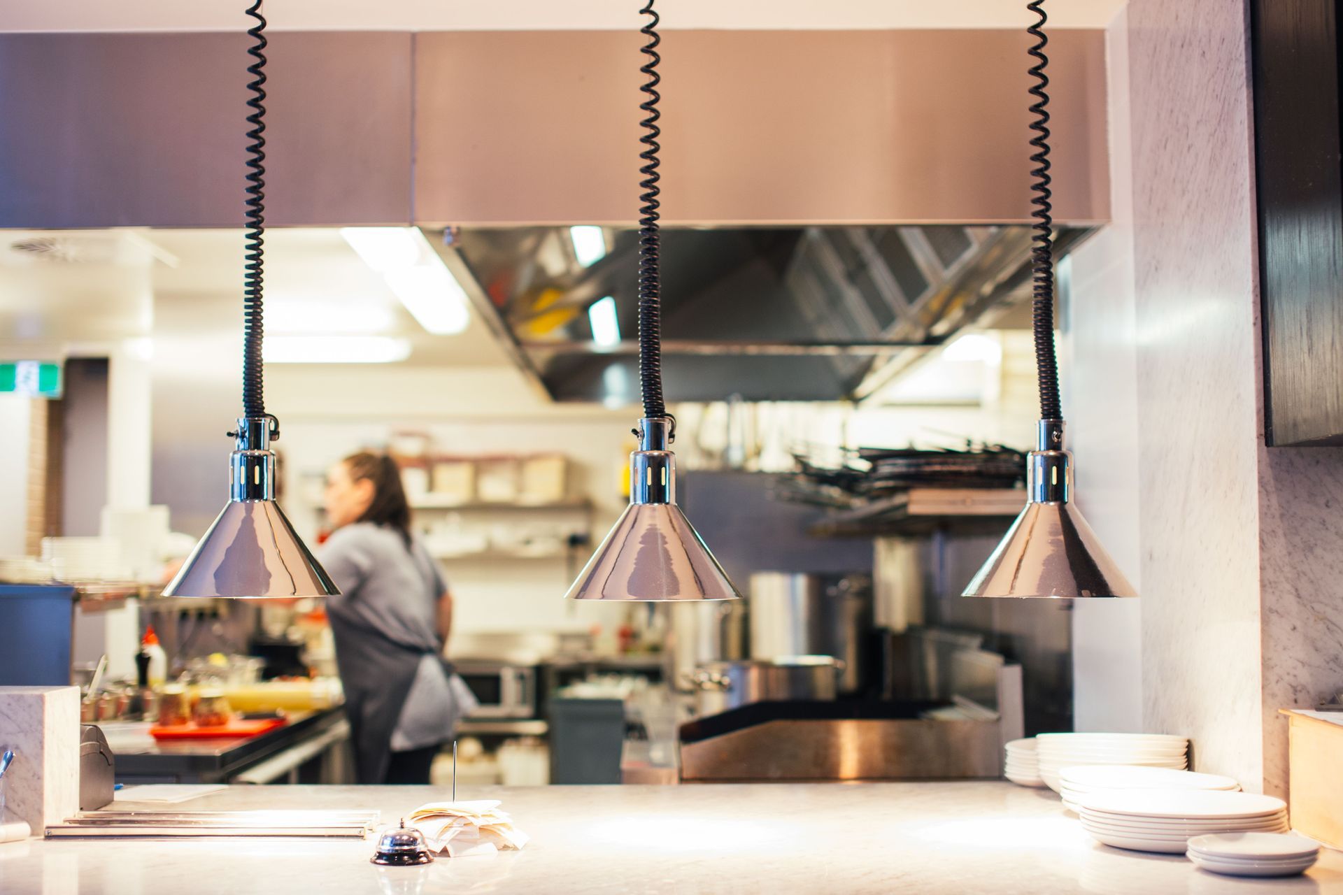 Modern restaurant kitchen with hanging heat lamps, a cook in the background, and plates on a counter
