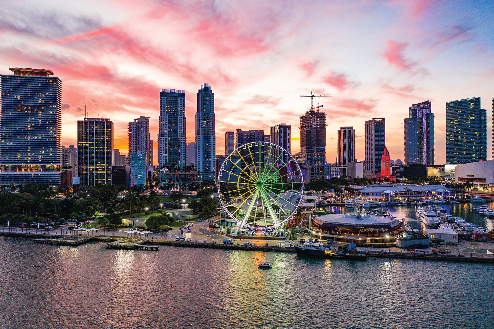Miami Downtown skyline at sunset viewed from the bay, featuring vibrant city lights, modern skyscrapers, and a Ferris wheel along the waterfront.