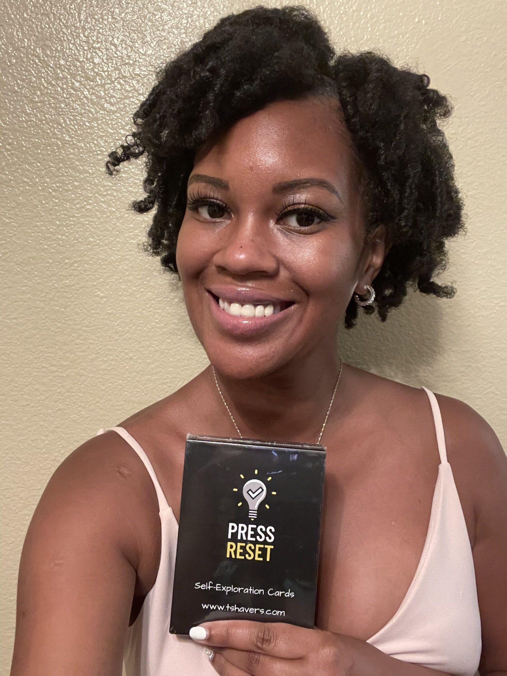 A pretty young woman stands against a wall and smiles while holding up her new Press Reset affirmation cards.