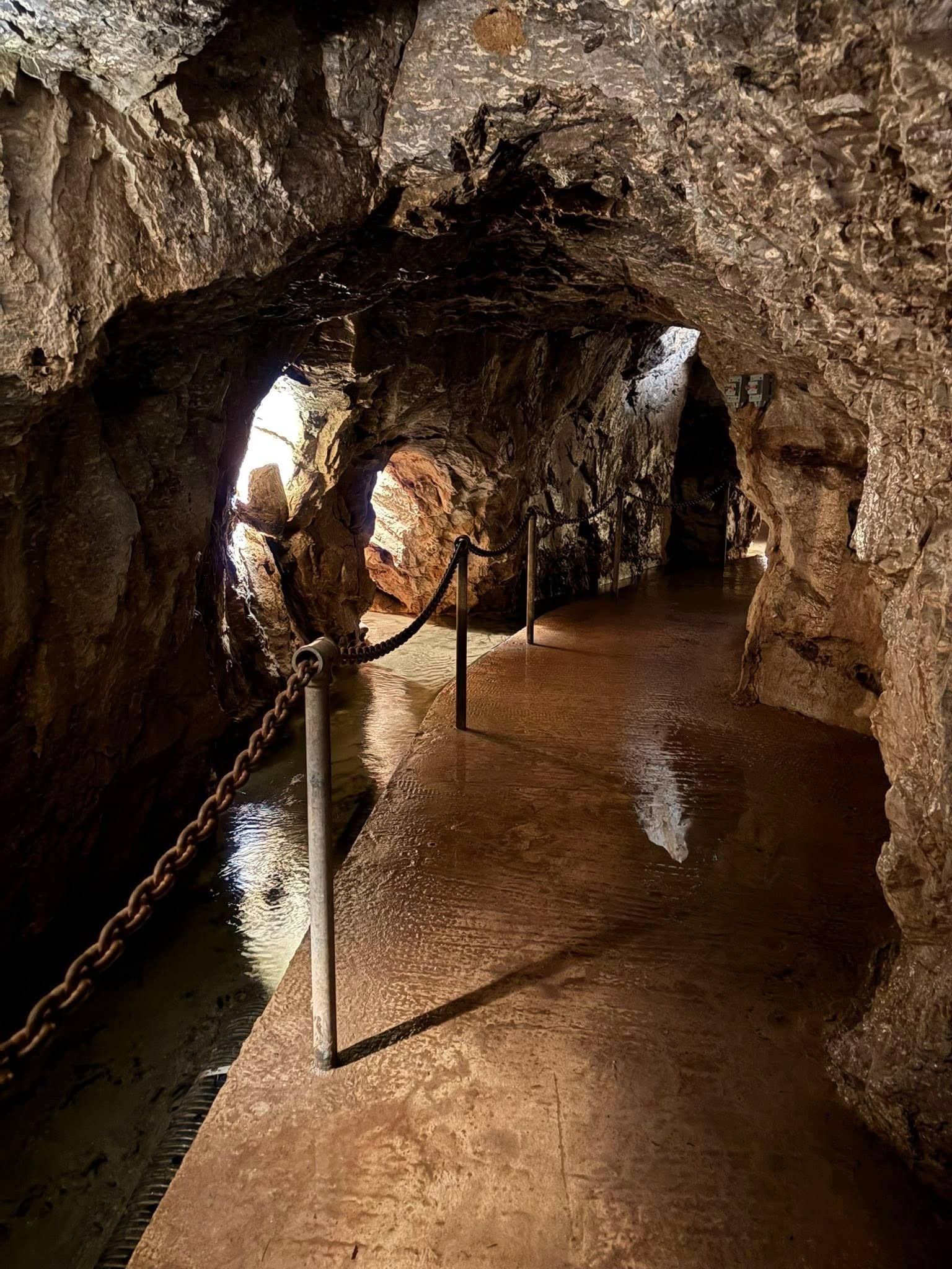 A narrow, wet cave passageway with a chained barrier, light at the end.