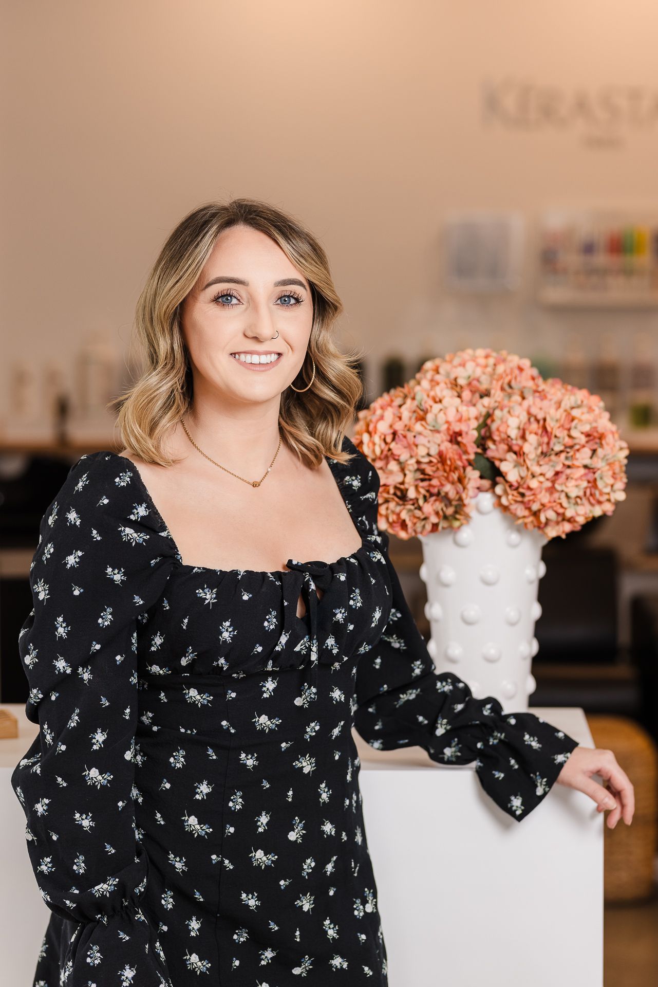 A woman in a black dress is standing in a salon next to a vase of flowers.