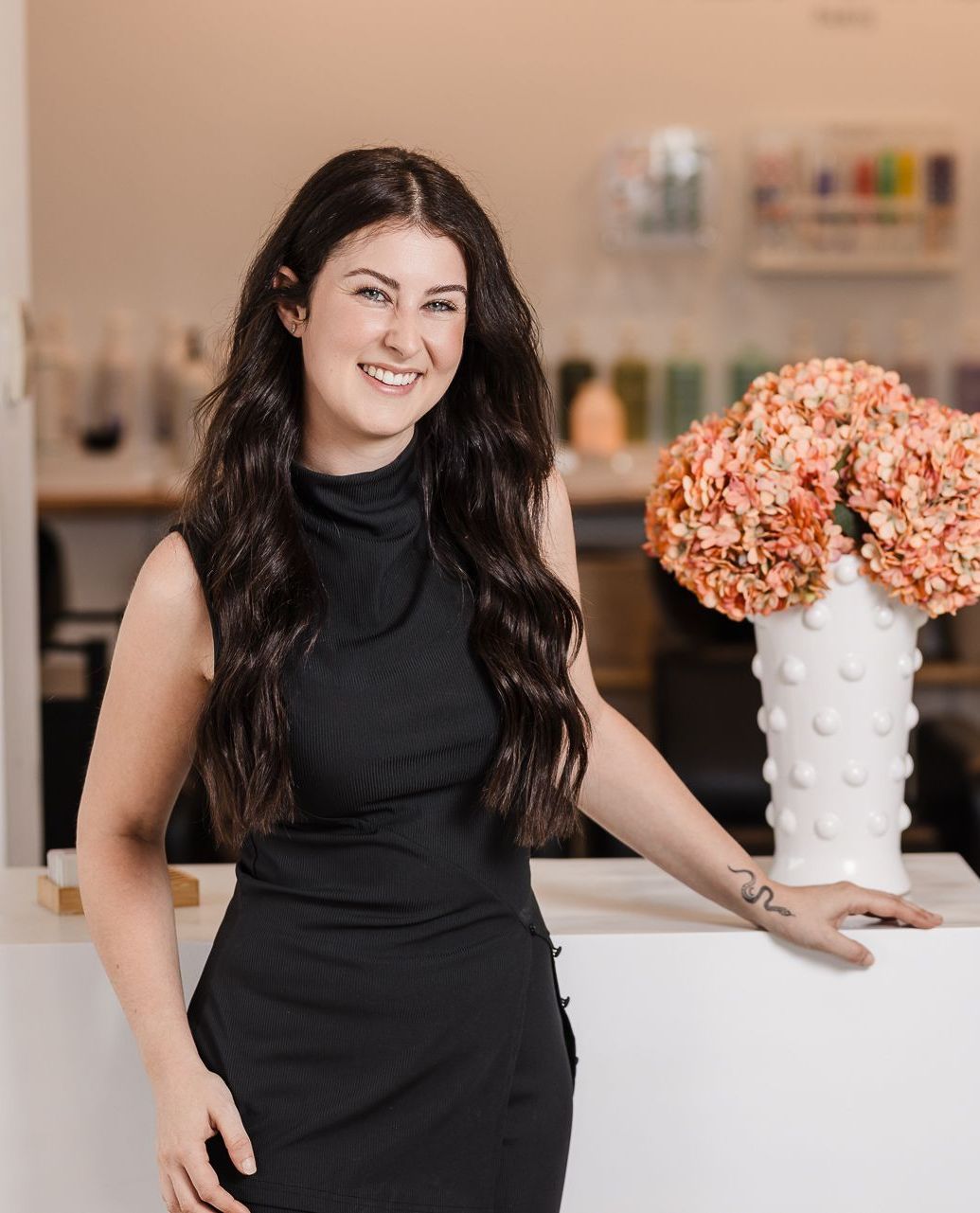 A woman in a black dress is standing in a salon next to a vase of flowers.