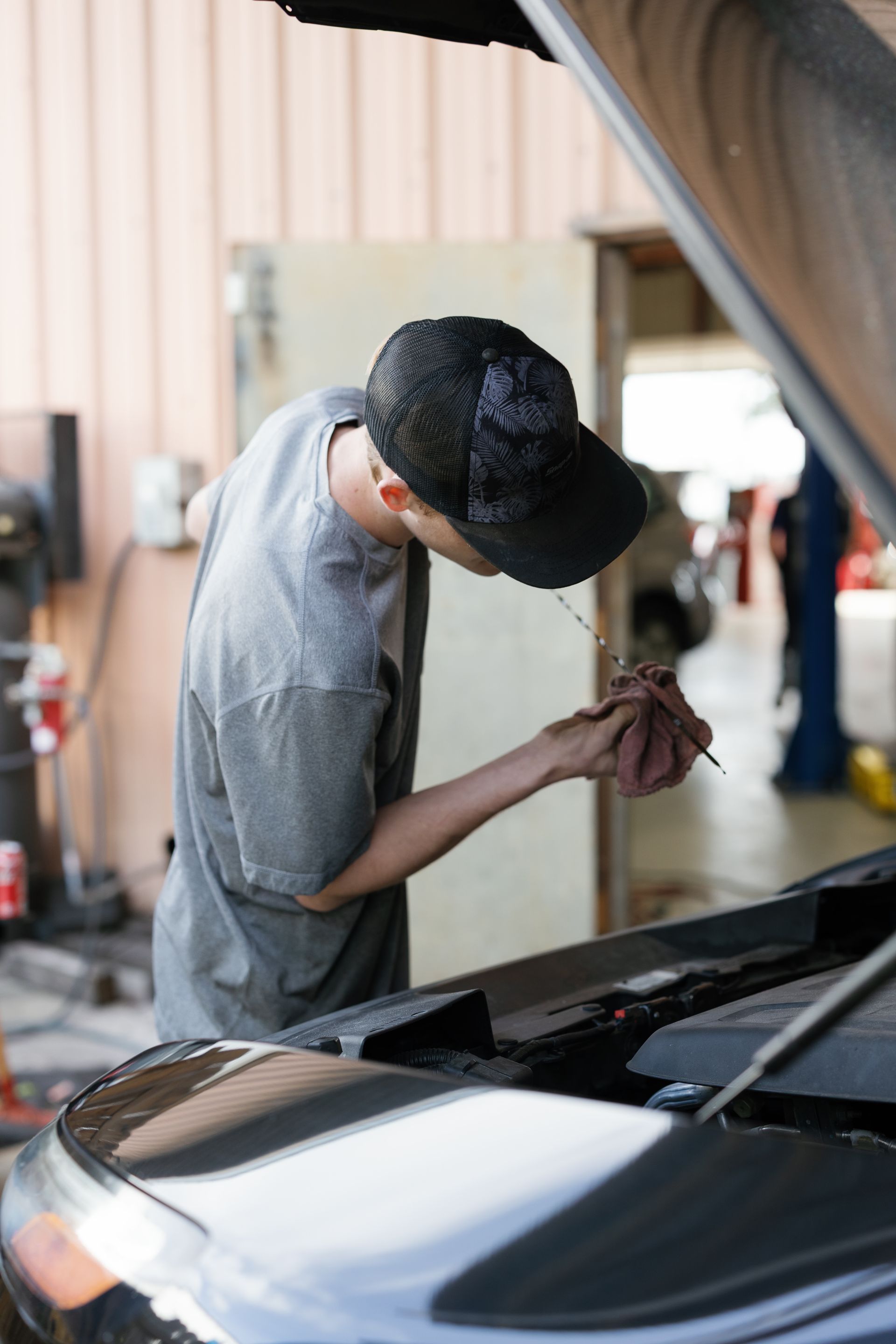 Mechanic checking car fluids with a dipstick in a garage; gray shirt, black cap.