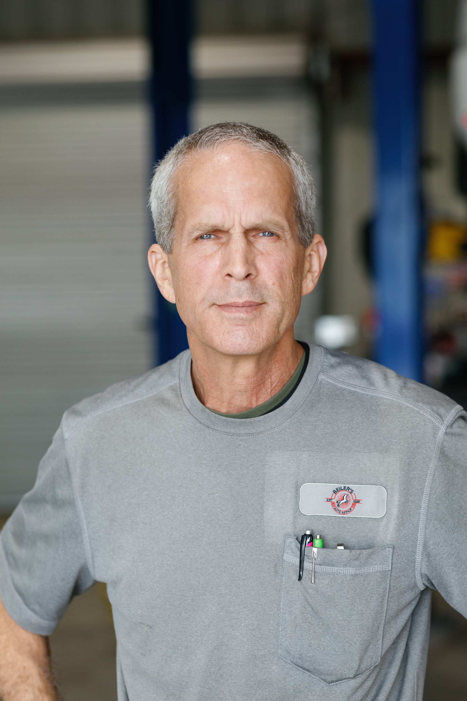 Man in gray shirt at a garage, looking at the camera. He has gray hair and a name tag.