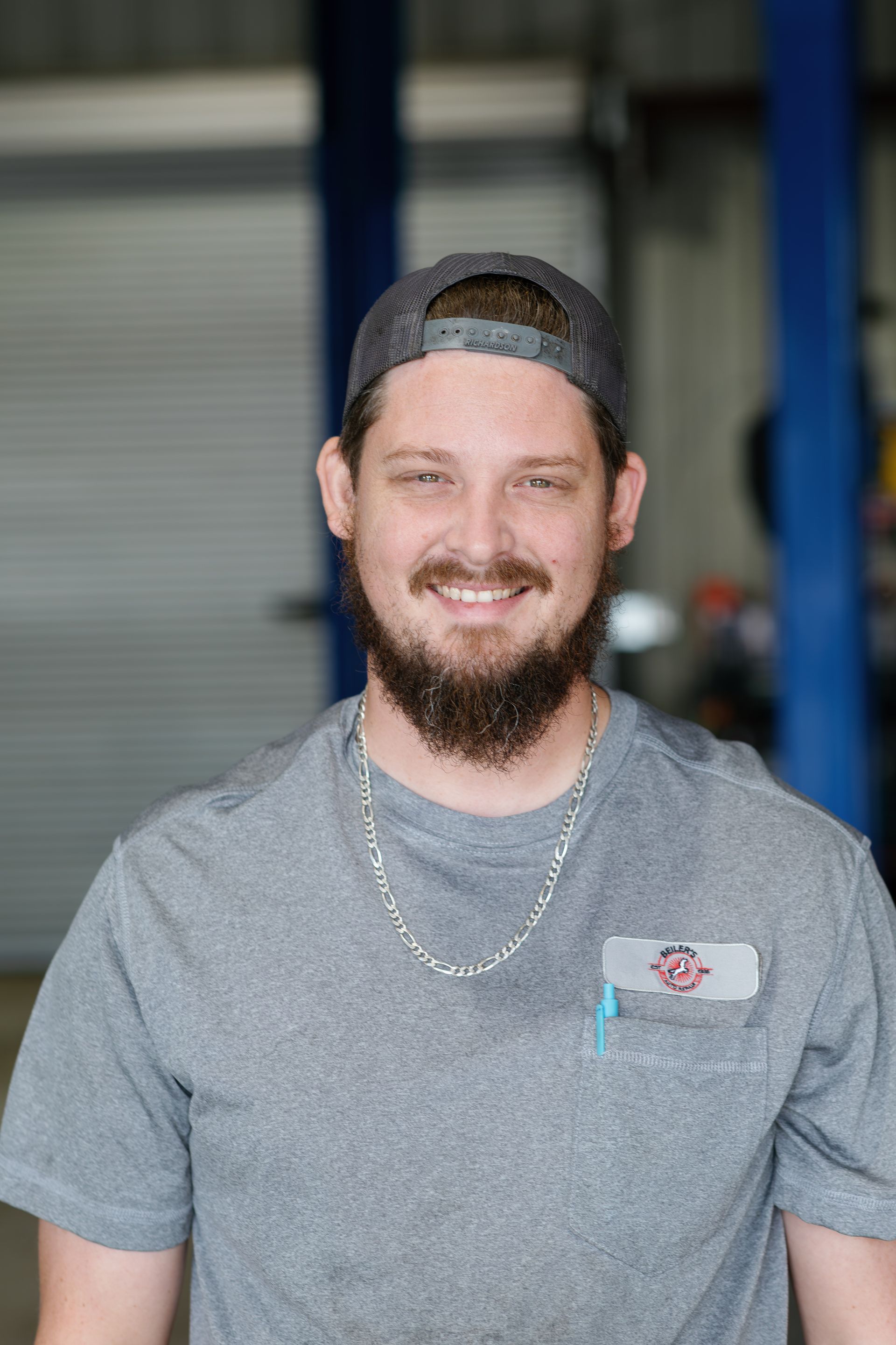 Man with a beard, wearing a grey t-shirt and hat, smiling in a garage setting.