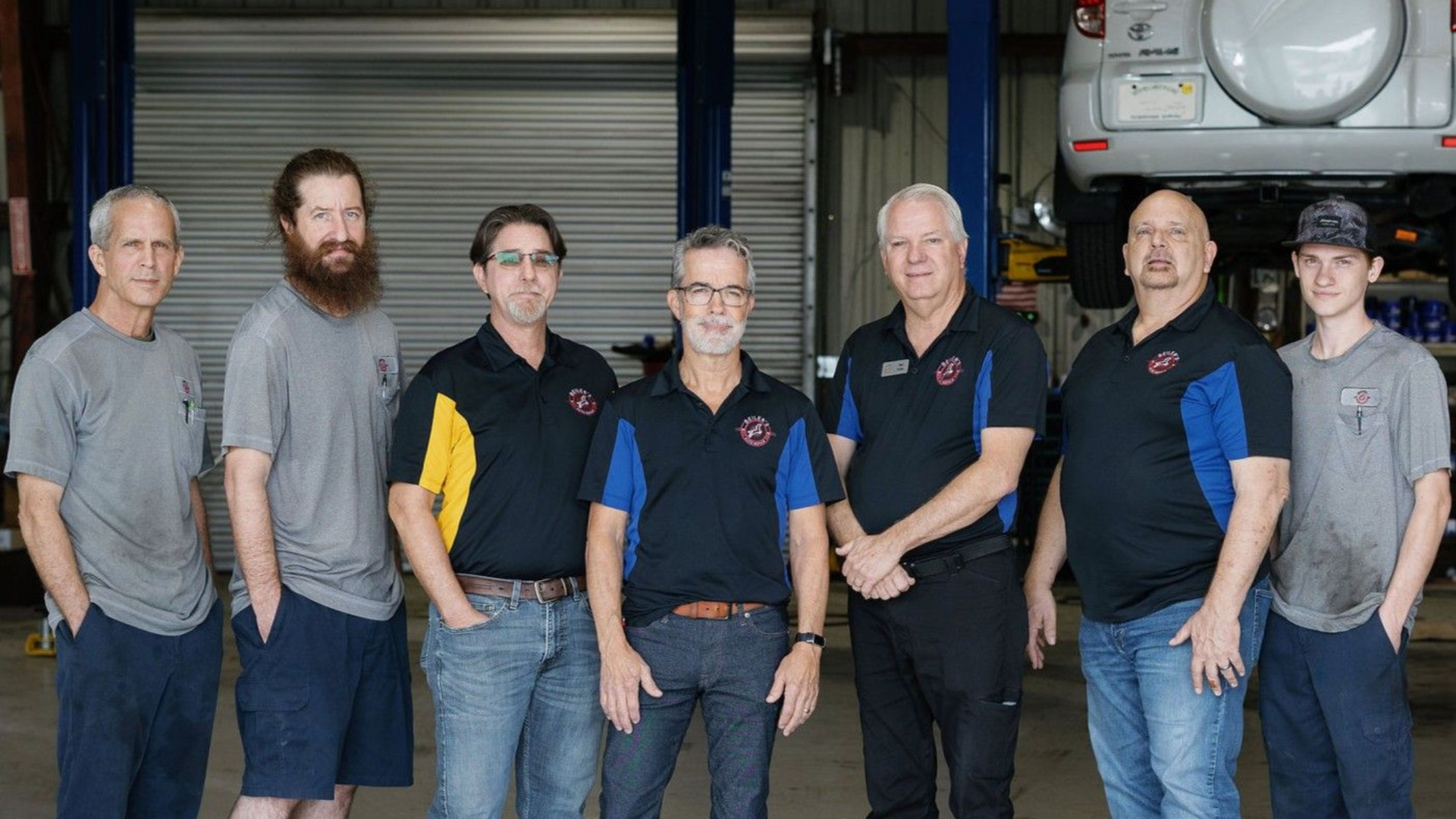 Group of eight auto mechanics posing in a garage with a car on a lift.