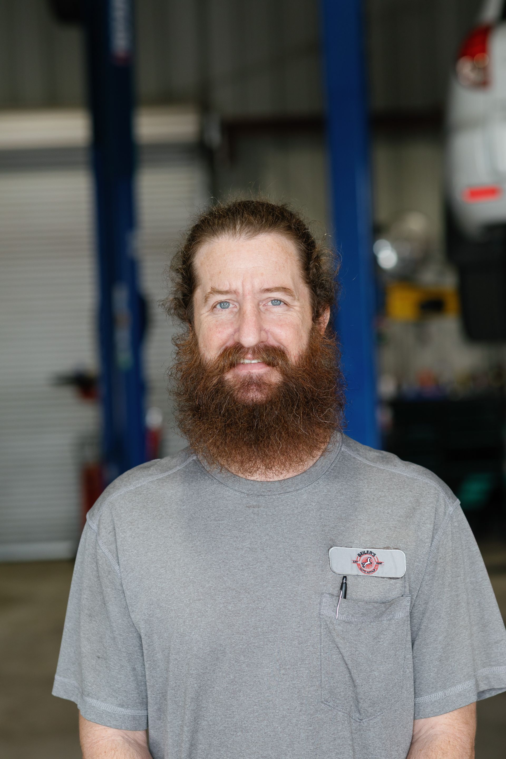 Man with a long, reddish-brown beard in a garage setting, wearing a gray shirt with a pocket.