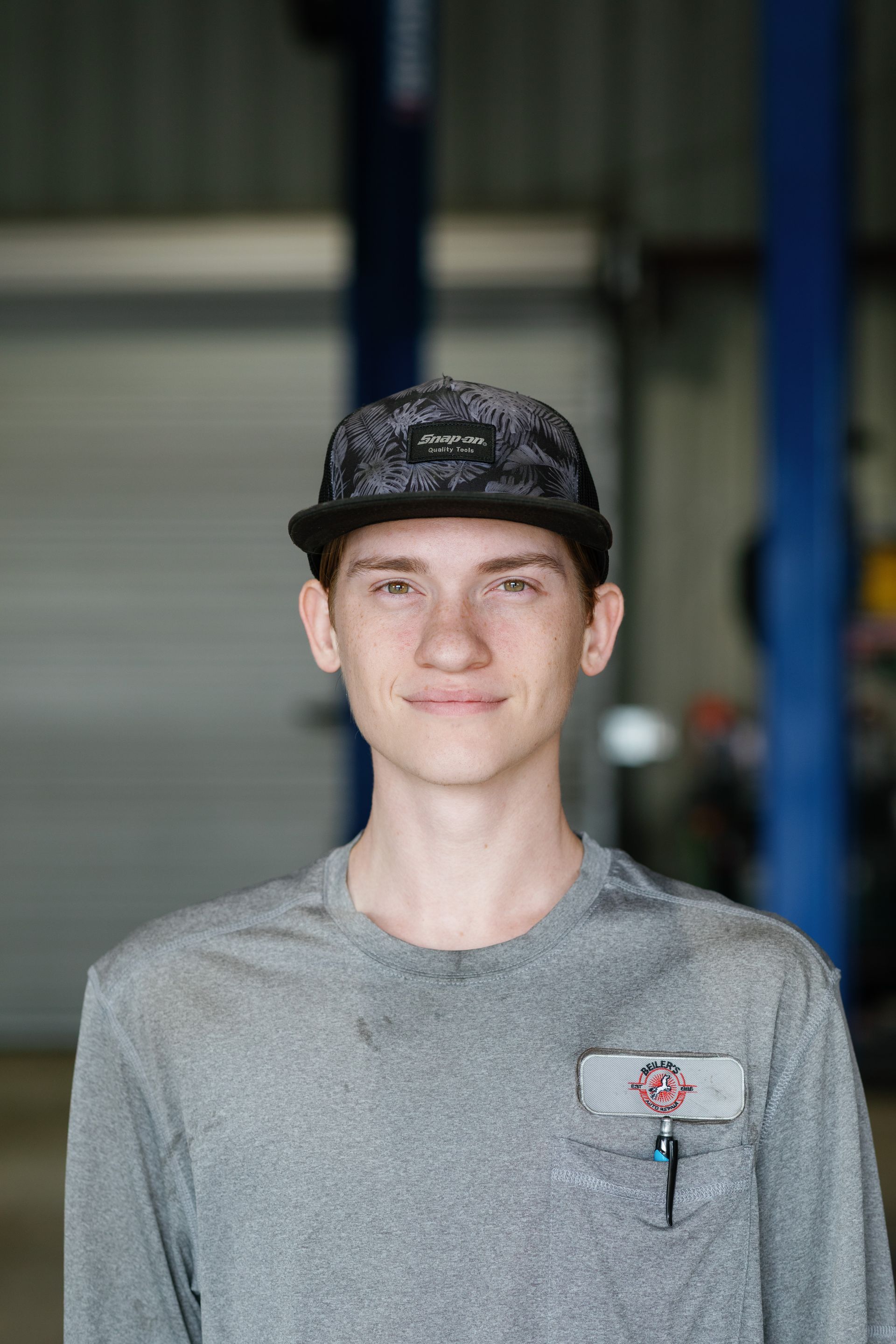 Young man in a cap and gray shirt smiles in a garage. He has freckles and a small pocket with a pin.
