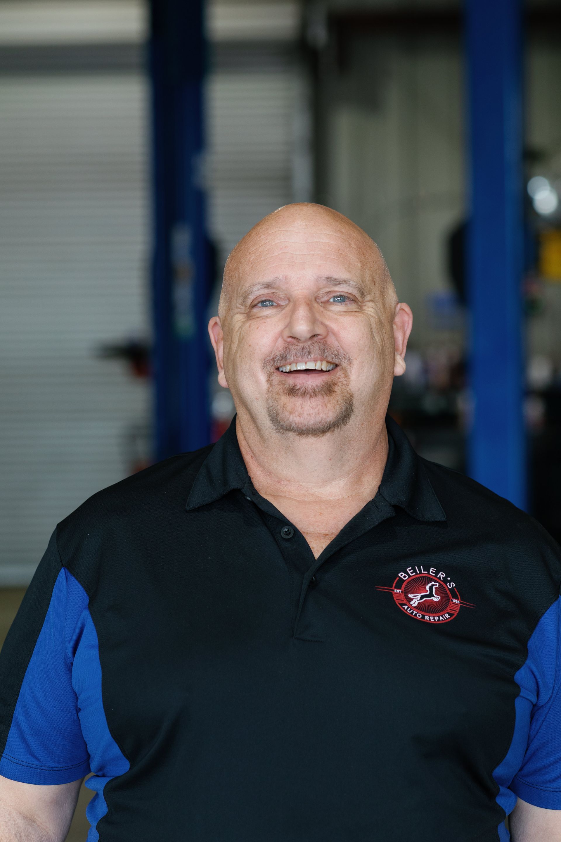 Bald man wearing a black and blue polo shirt smiles in a garage setting.
