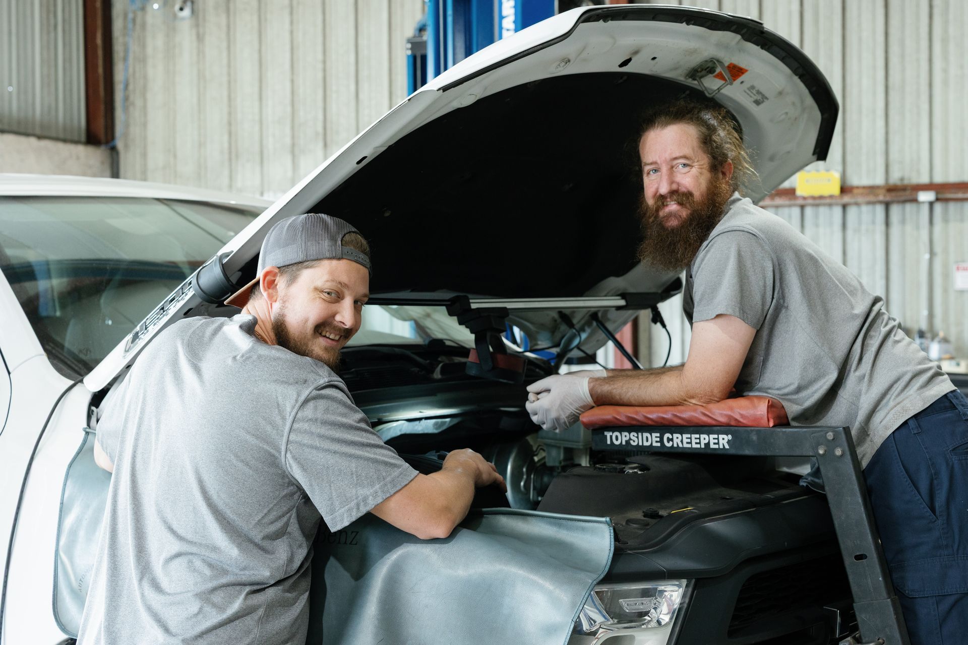 Two mechanics smiling, working on a car engine in a garage.
