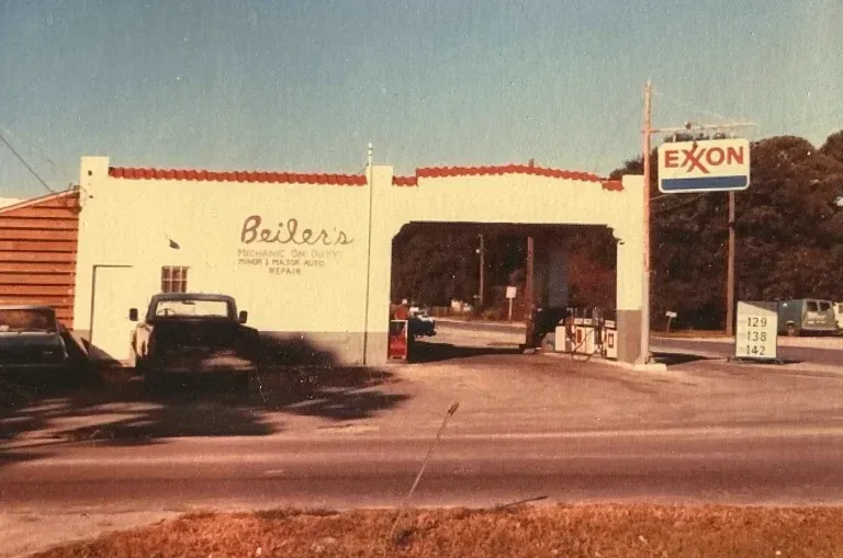 Gas station, Beiler's, with an Exxon sign. A white building with a drive-through under an archway. A truck and car parked outside.