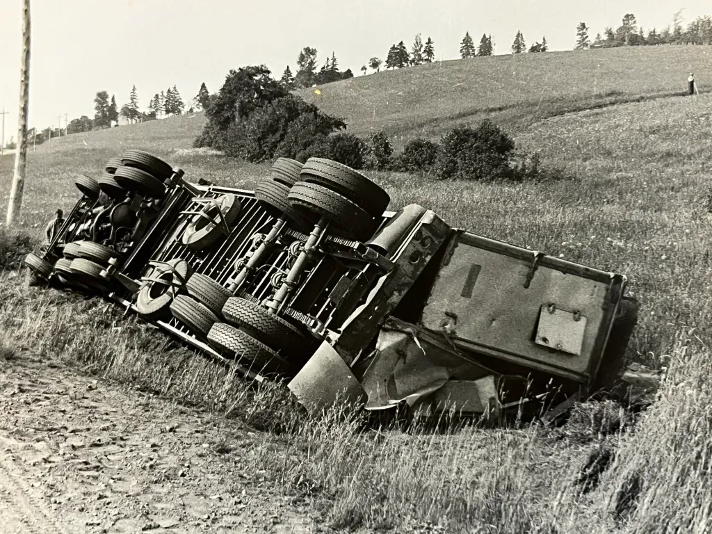 Overturned semi-truck on its side in a grassy ditch next to a road, rolling hills in the background.