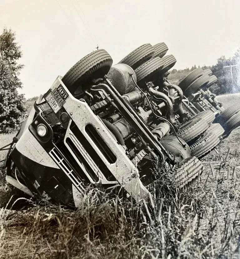 Semi-truck overturned in grassy roadside ditch. Visible tires, chassis, and cab.