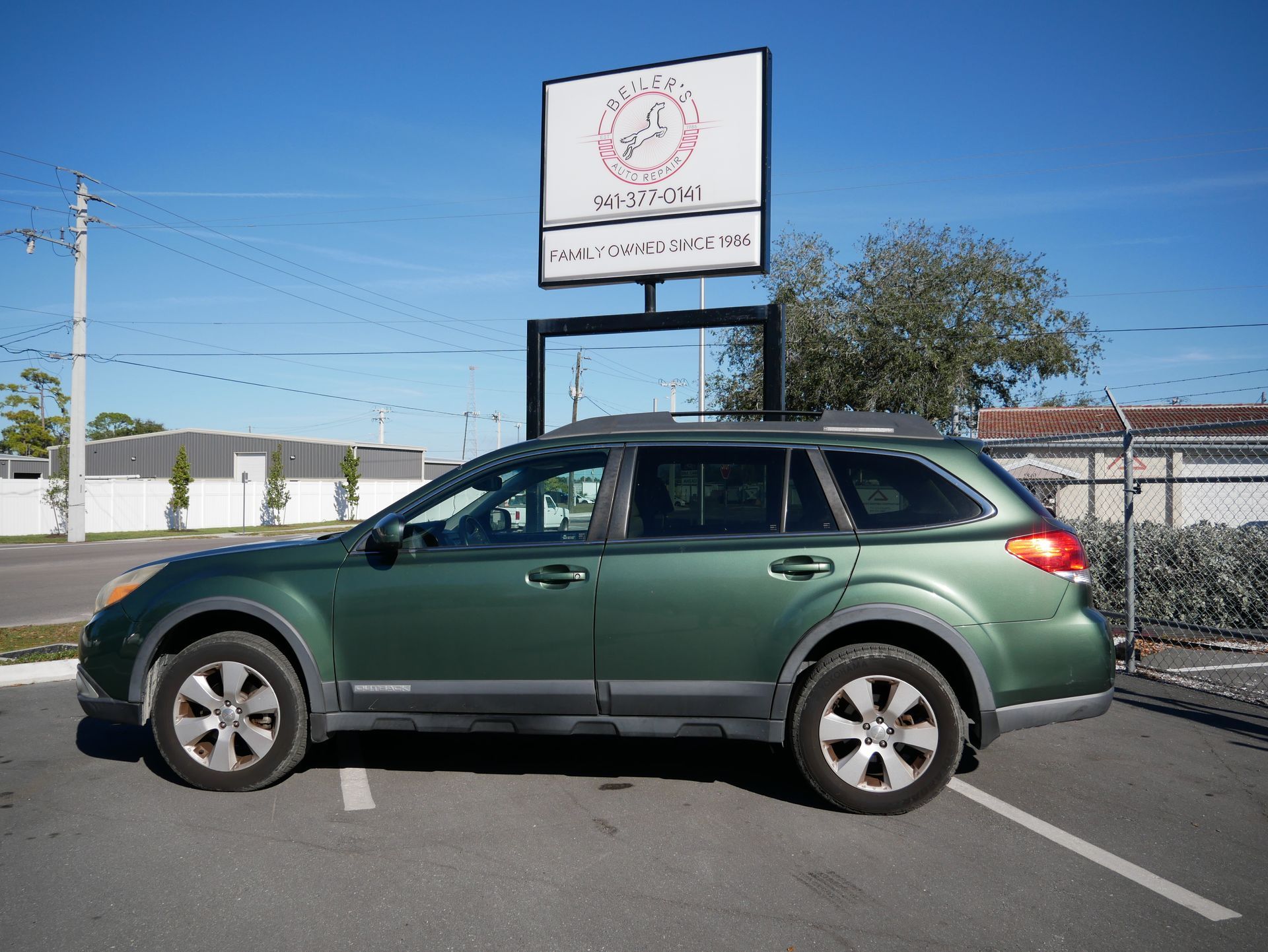 Green Subaru Outback parked in a lot, with a sign in the background under a blue sky.
