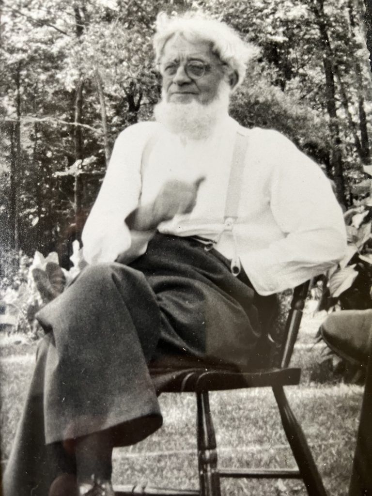 Older person with white hair and beard, wearing suspenders, sits outdoors in a chair, gesturing with his hand.