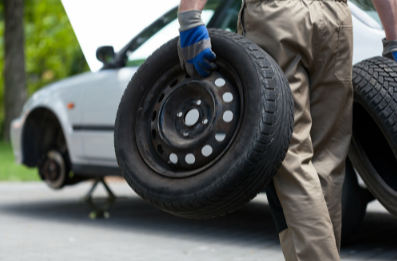 Picture of a man holding two tires walking toward a vehicle that is hoisted up with no tires on it.