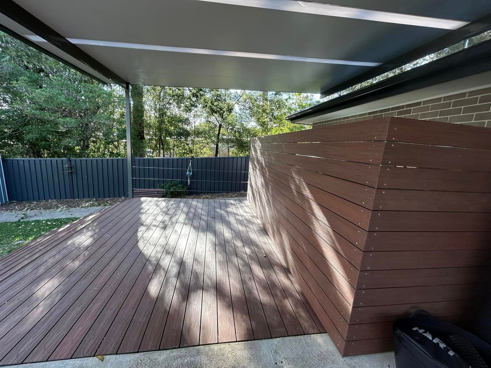 Brown pergola over a patio, casting striped shadows on the stone. Green lawn, trees, and blue sky.