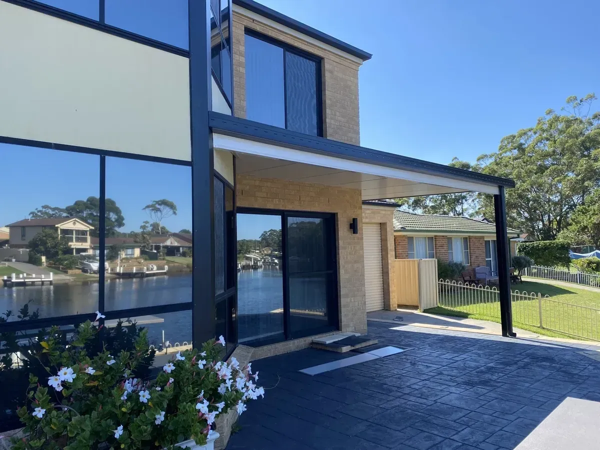 Modern two-story home with large windows, black awning, and waterfront view on a sunny day.