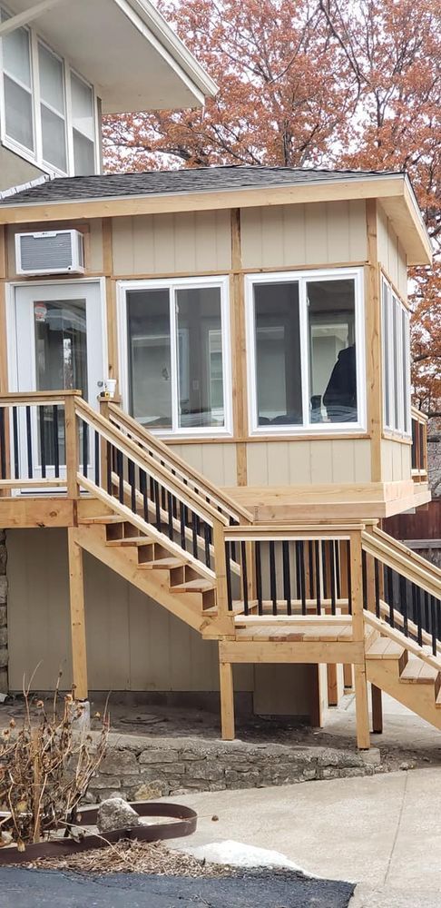 A light-colored sunroom atop a deck with stairs, near a building and trees.