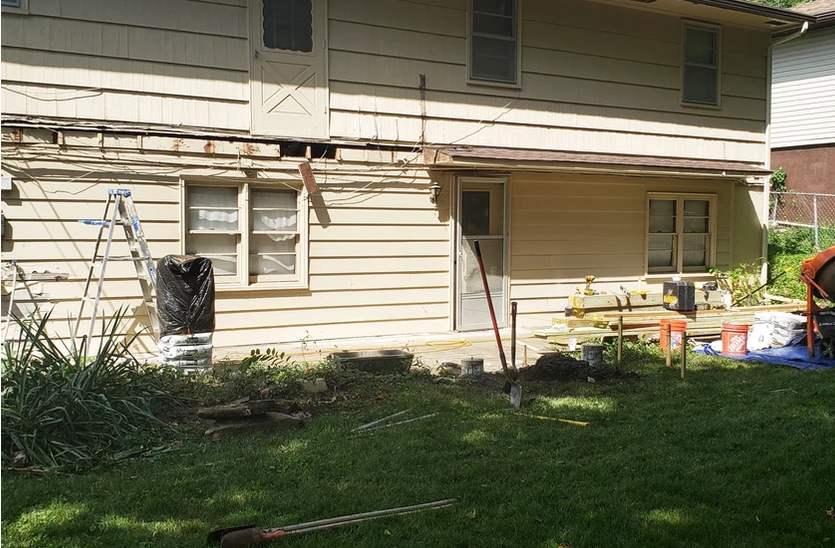 Backyard of a house under renovation; exposed wood, ladder, trash, and materials on grass.