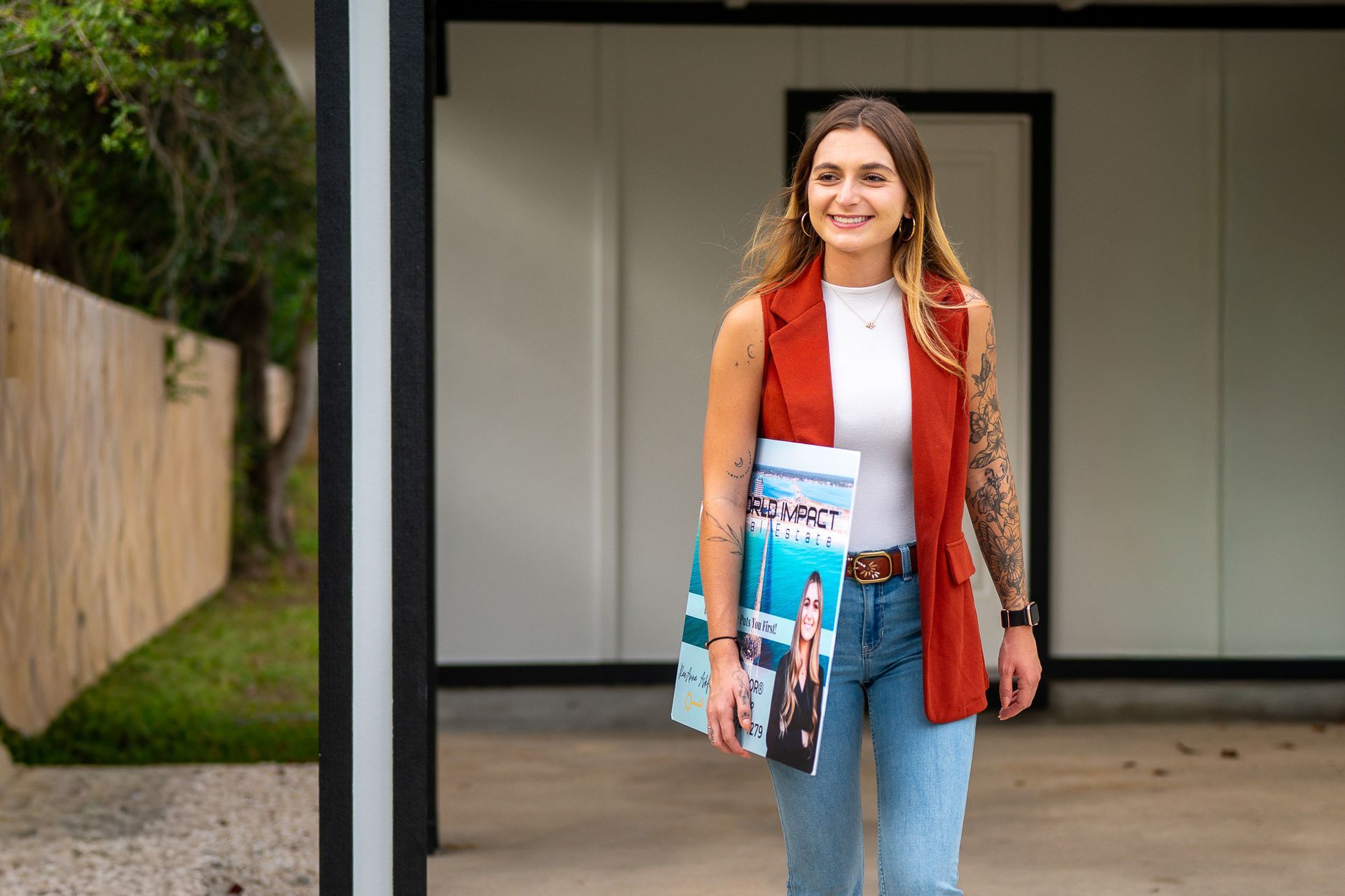 Woman with arm tattoos, wearing a rust vest, jeans, and holding a magazine, standing outside a modern building.