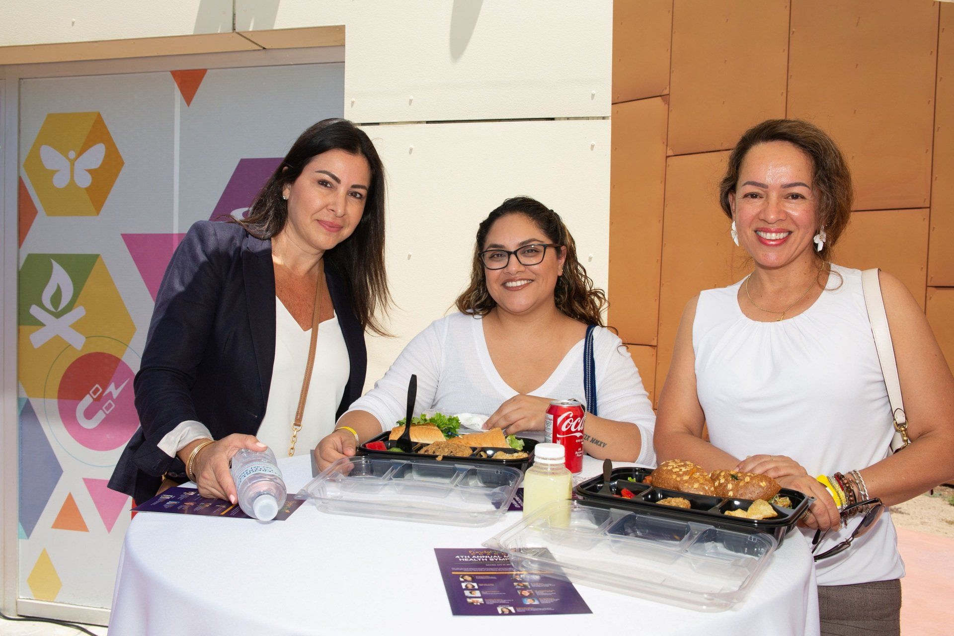 Three women are standing around a table with food and drinks.