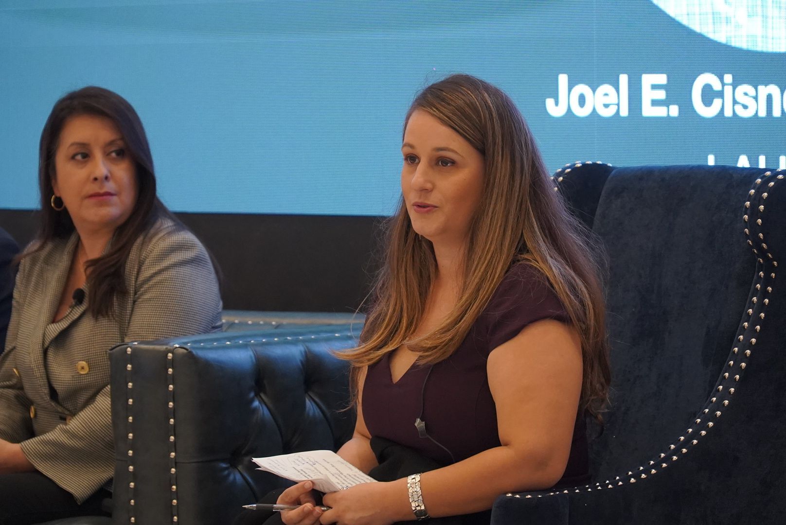 Two women are sitting on a couch in front of a screen that says joel e. cisner