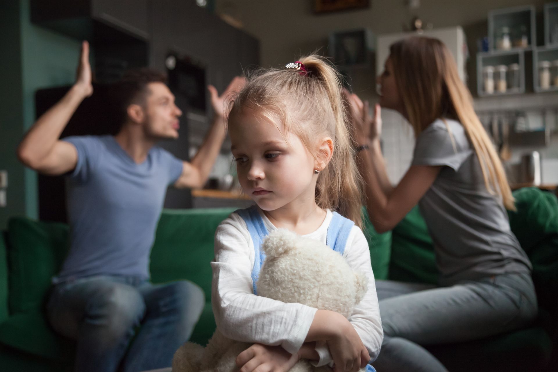 A little girl is holding a teddy bear while her parents argue in the background.