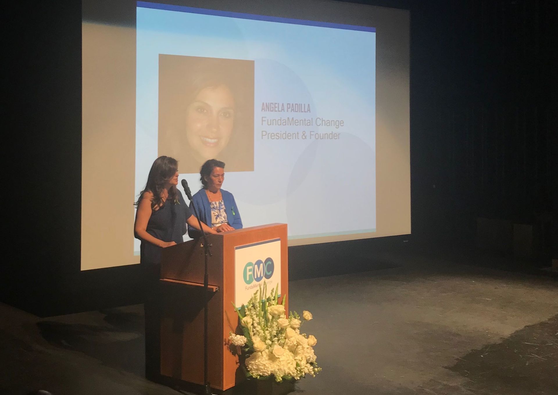 Two women are standing at a podium in front of a large screen.
