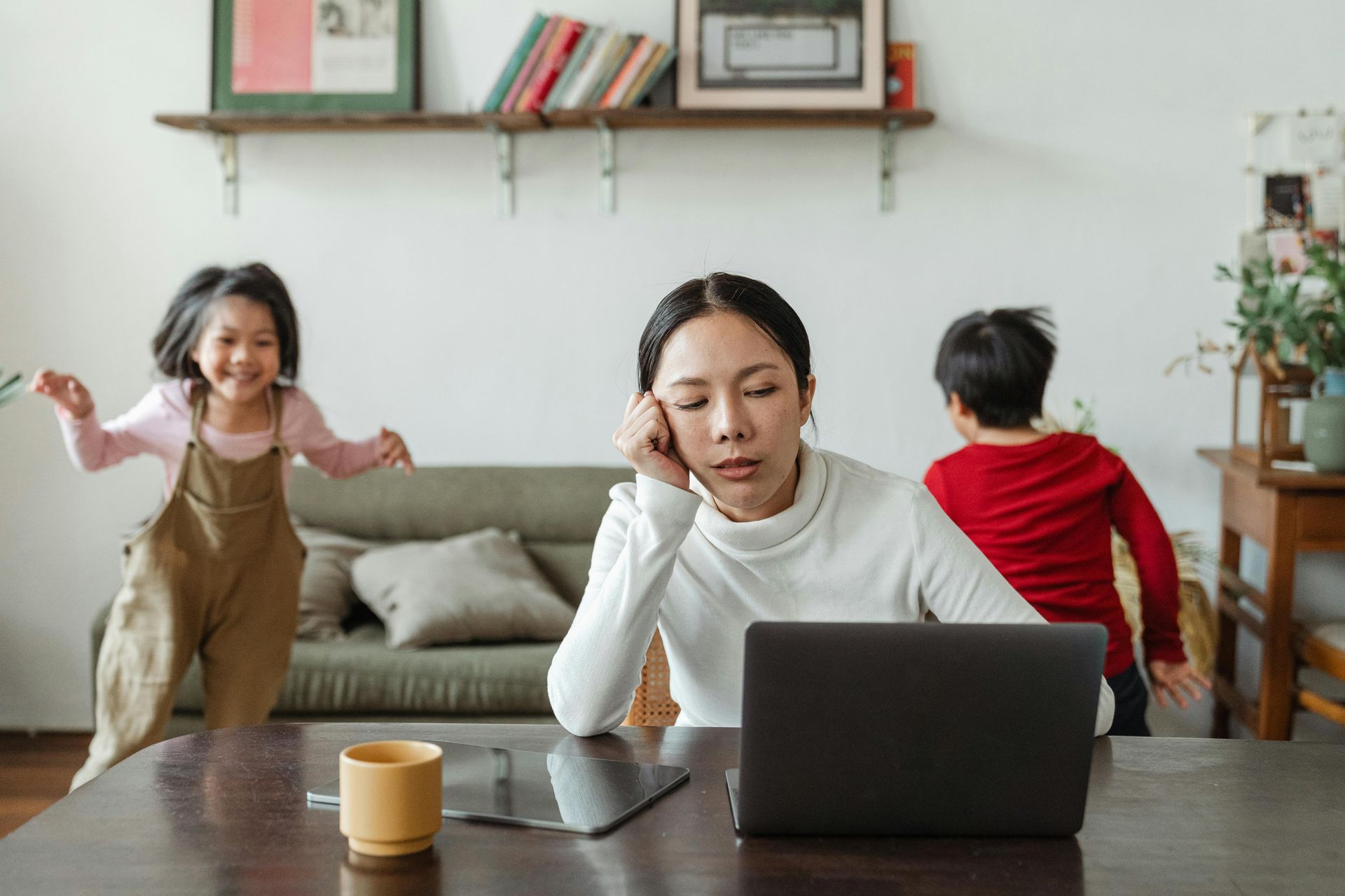 A woman is sitting at a table using a laptop computer while two children play in the background.