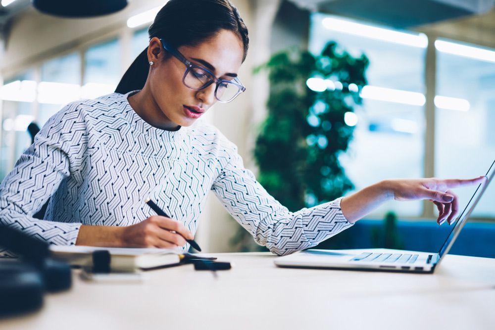 A Woman is Sitting at a Desk Using a Laptop Computer and Writing in a Notebook — Rauseo Group in Zuccoli, NT