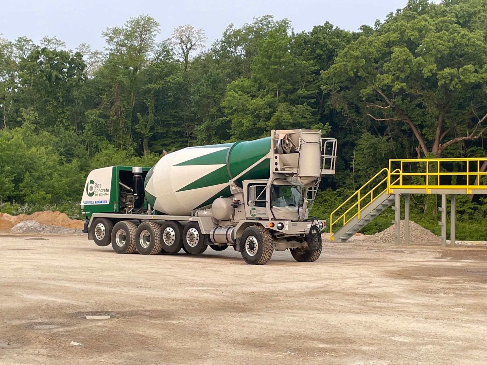 Cement truck, white and green, on a gravel lot near a forested area and a yellow platform.