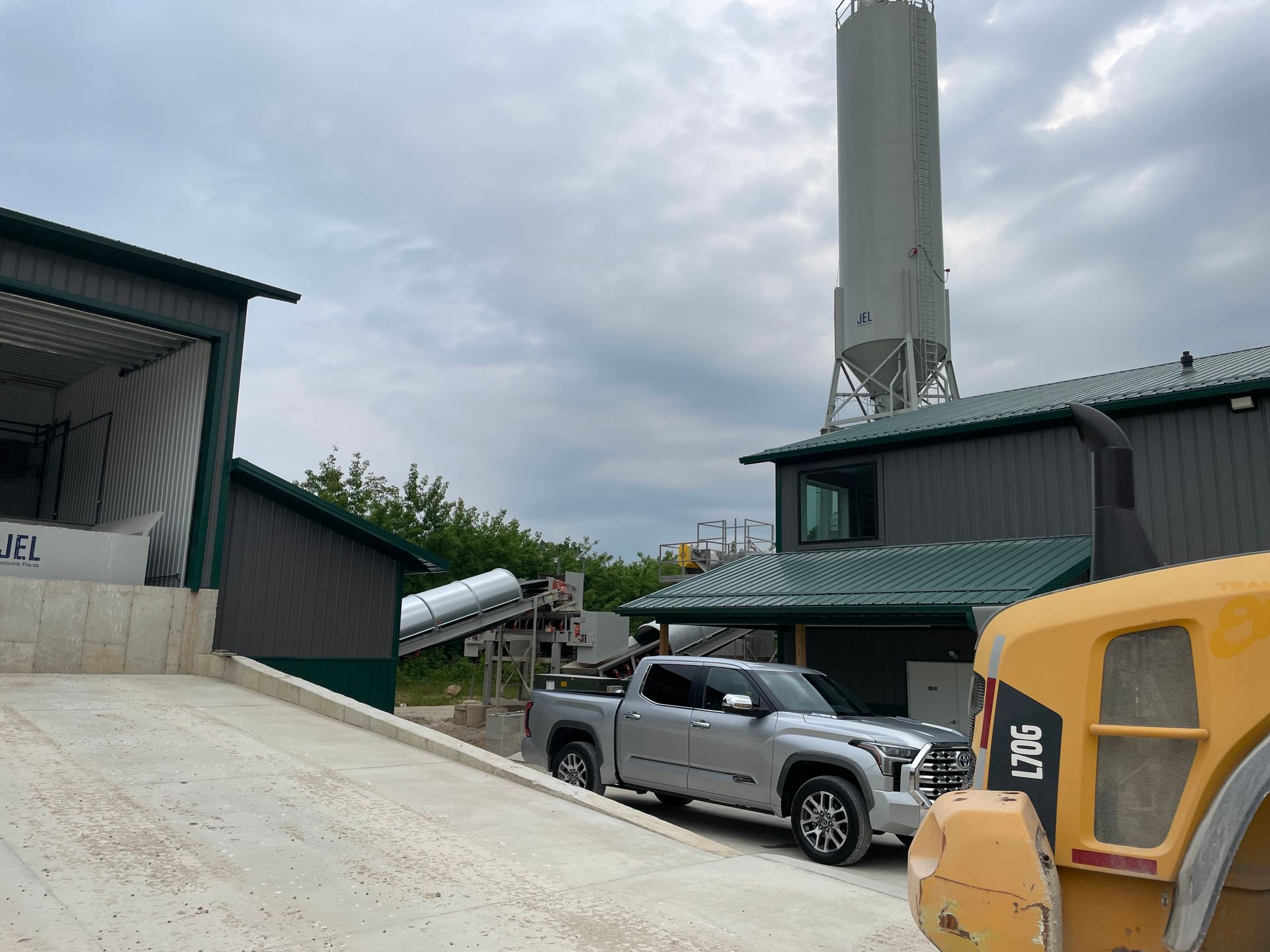 Silver truck parked near industrial building with tall silo, under a cloudy sky.