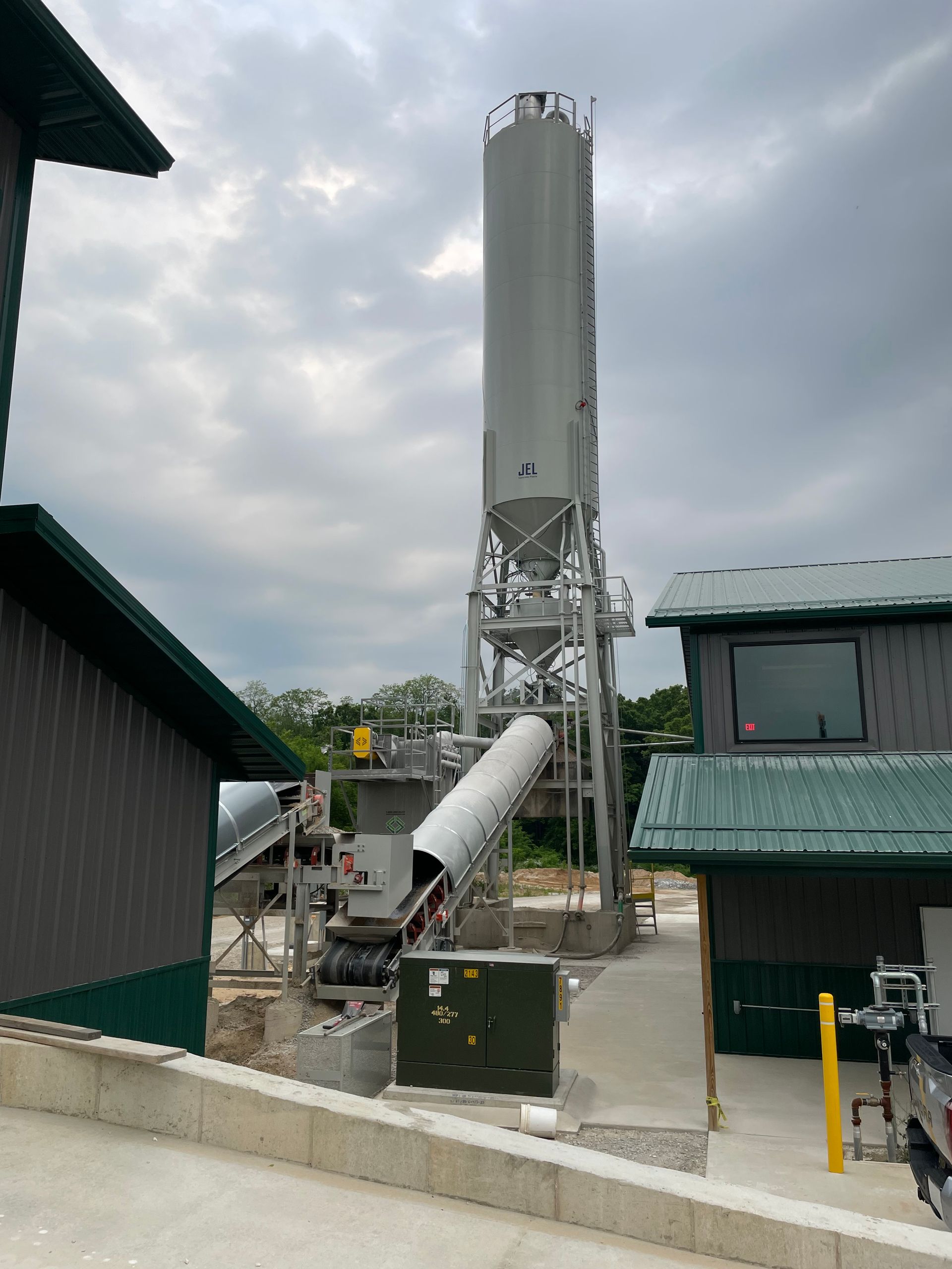 Concrete plant with tall silo, conveyor, and machinery between two buildings under cloudy sky.
