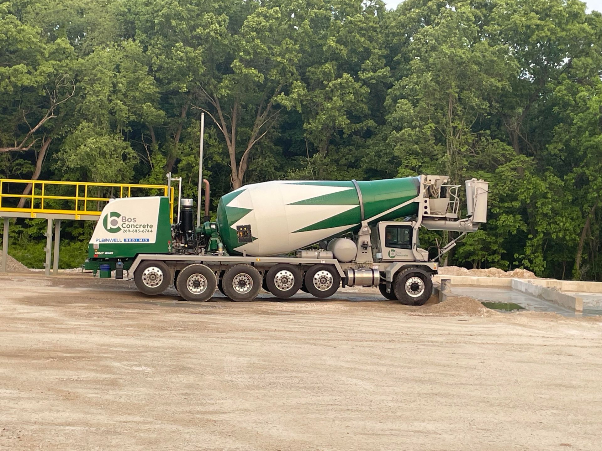 Cement truck, green and white, parked on gravel, with trees in the background.