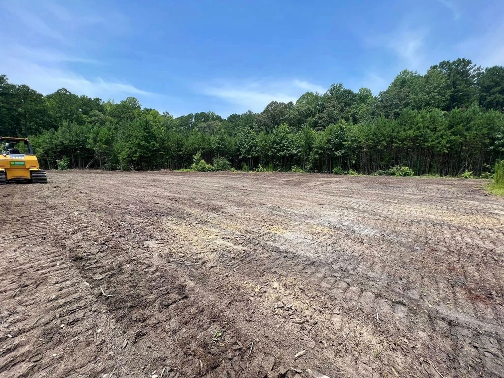 A yellow bulldozer is working on a dirt field with trees in the background.