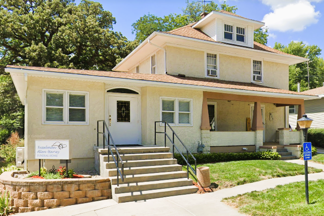 Two-story cream-colored building with porch, stairs, and a sign. A tree is in the background.