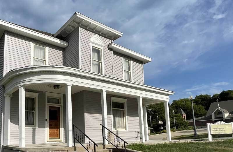 Two-story gray house with white trim and a porch. Cloudy sky background, church visible in the distance.