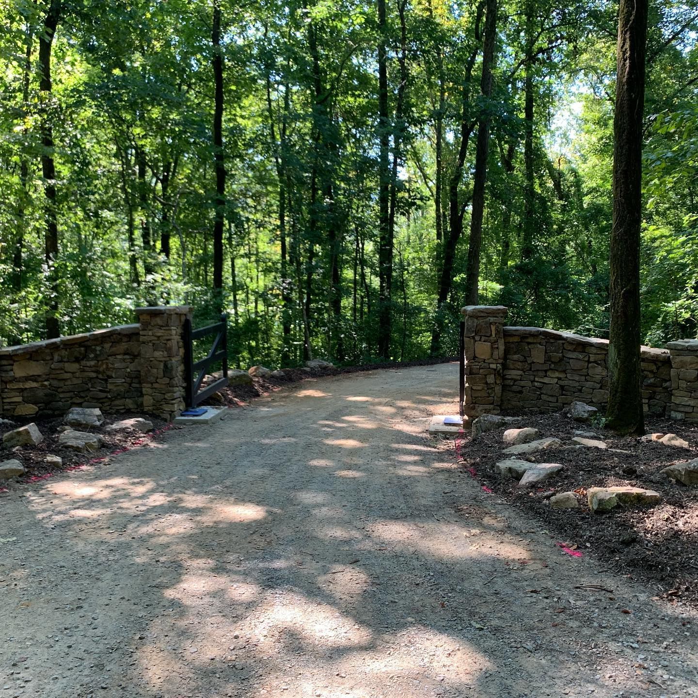 A gravel road going through a forest with trees and a stone wall.