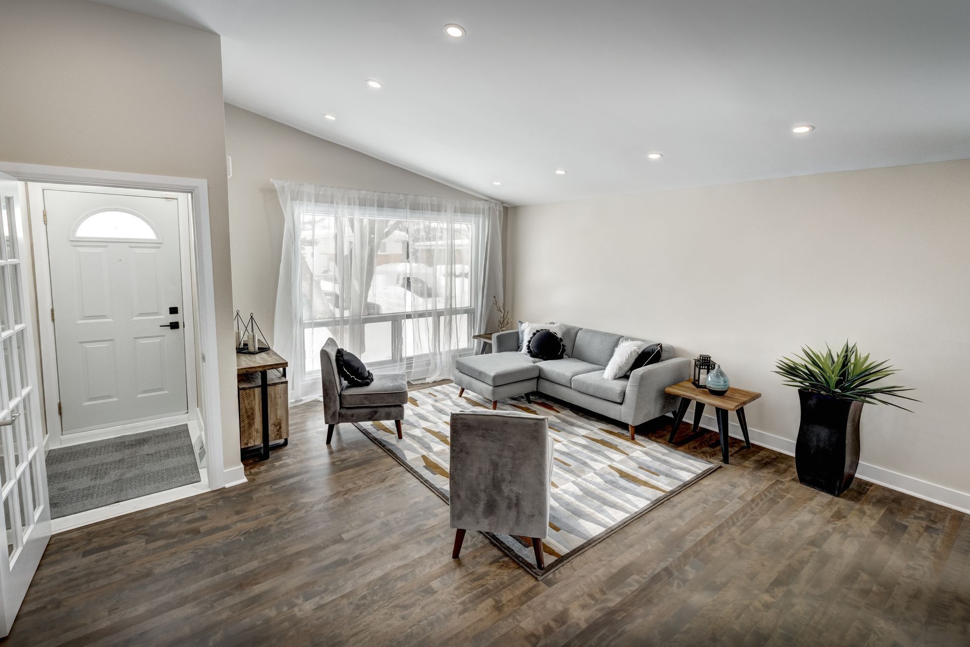 Living room with gray sofa, armchairs, and wood floor. Large window with sheer curtains.