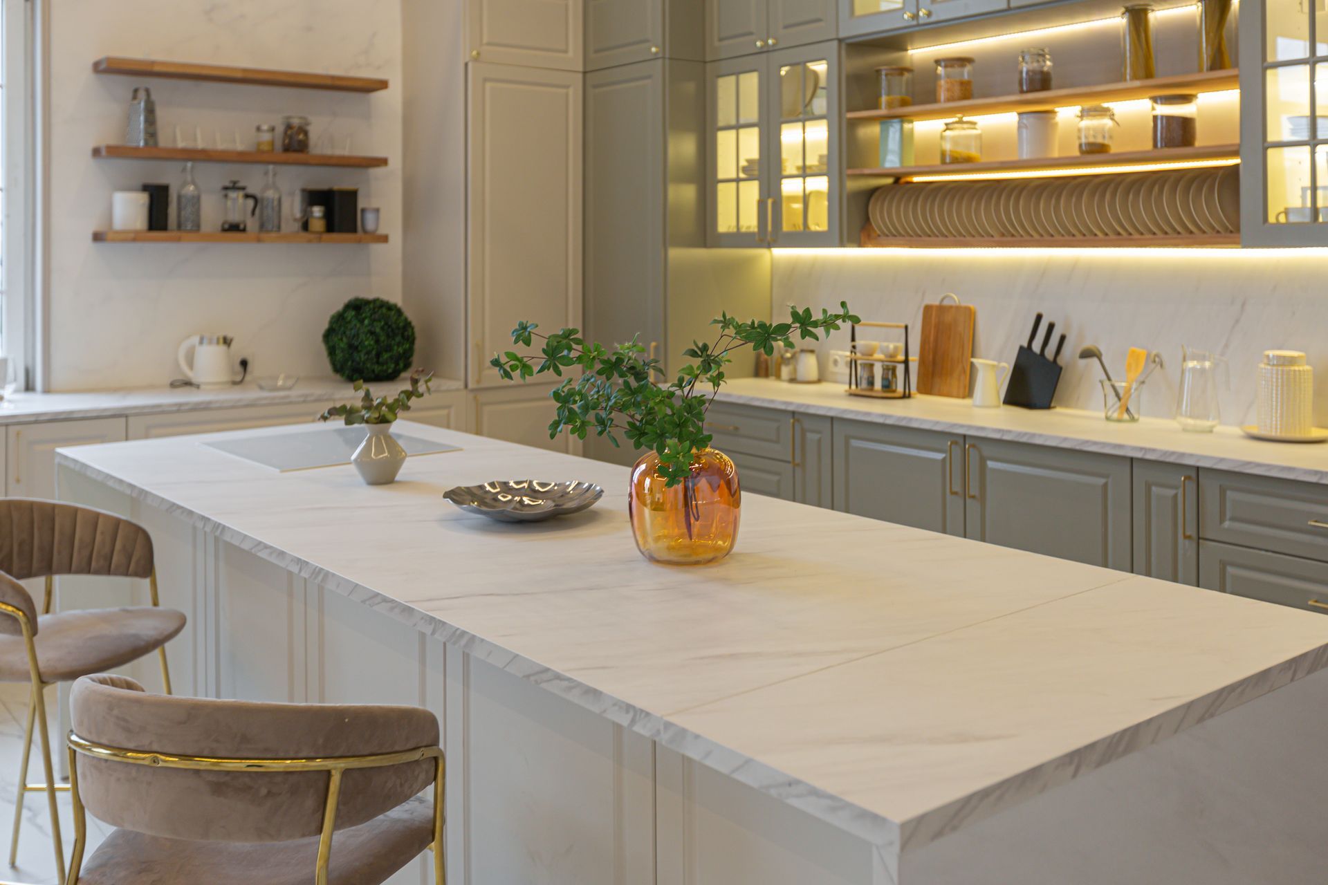 Elegant kitchen with marble island, wooden shelves, green cabinets, and gold accents.