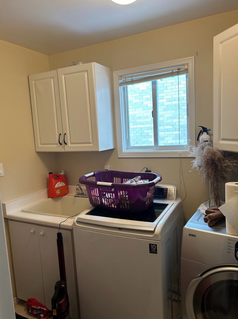Laundry room: white cabinets, sink, washing machine, basket of clothes, window.