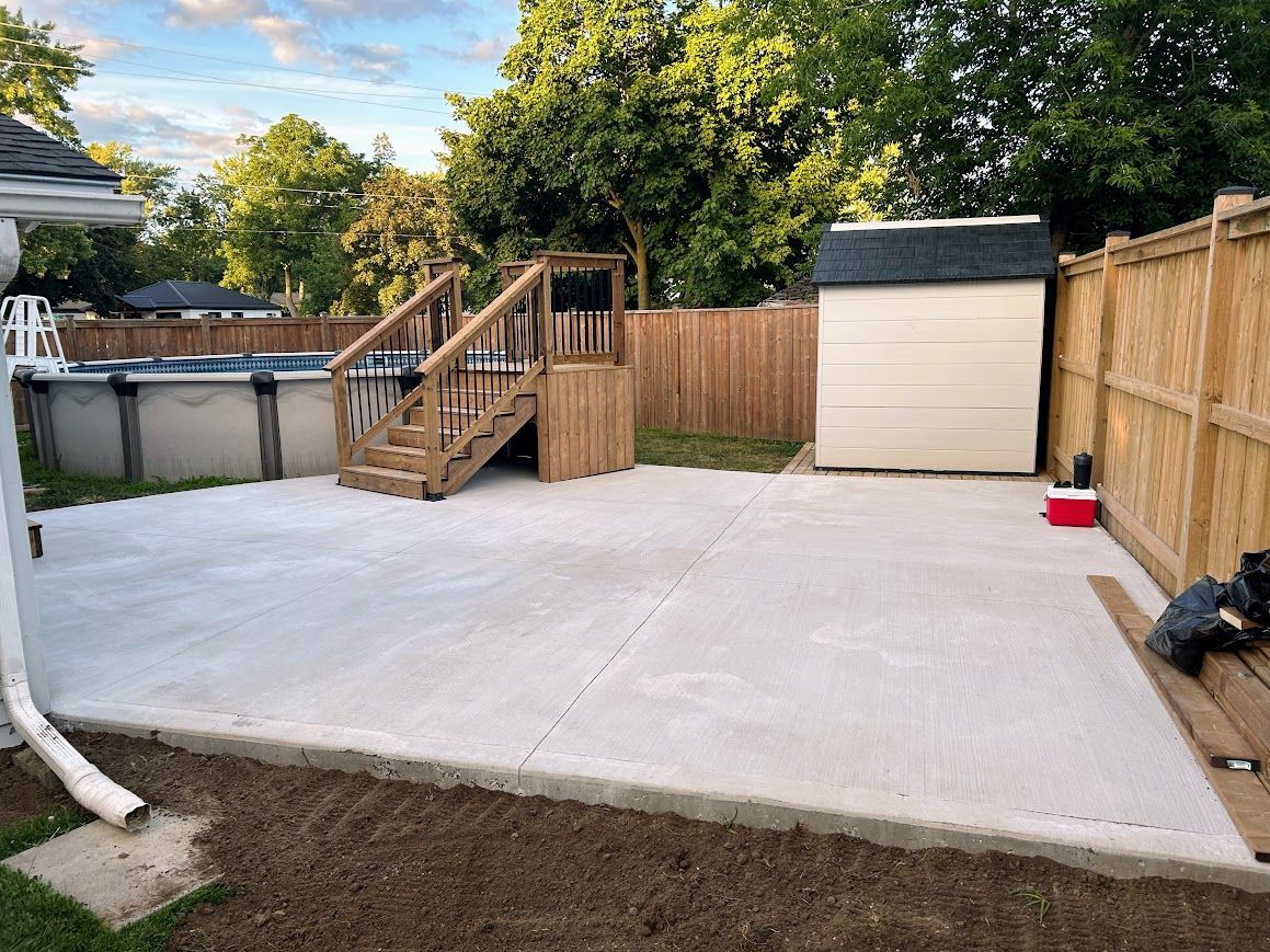 Concrete patio with pool, play structure, shed, and fence in a backyard.