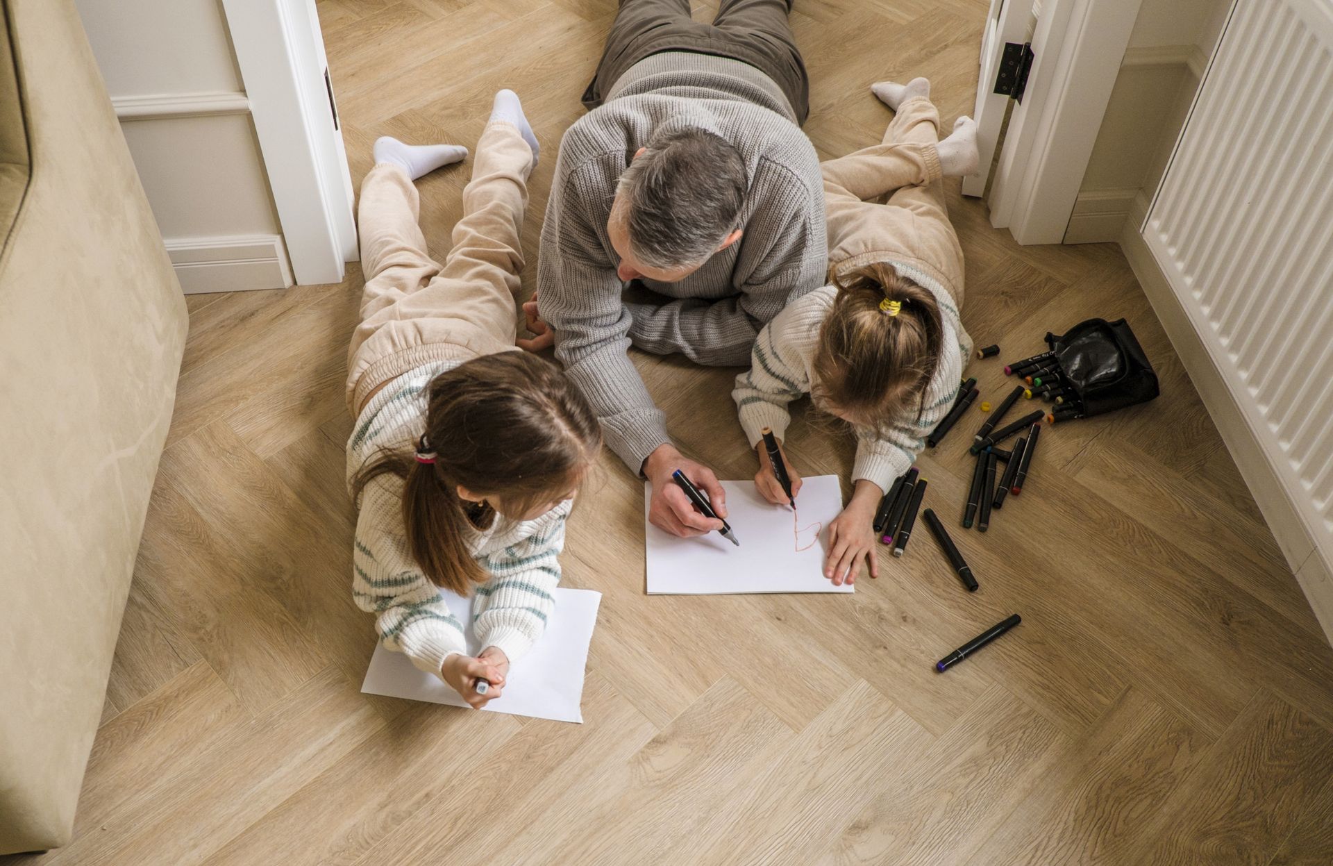 Man drawing with two children on a wooden floor; pens and paper.