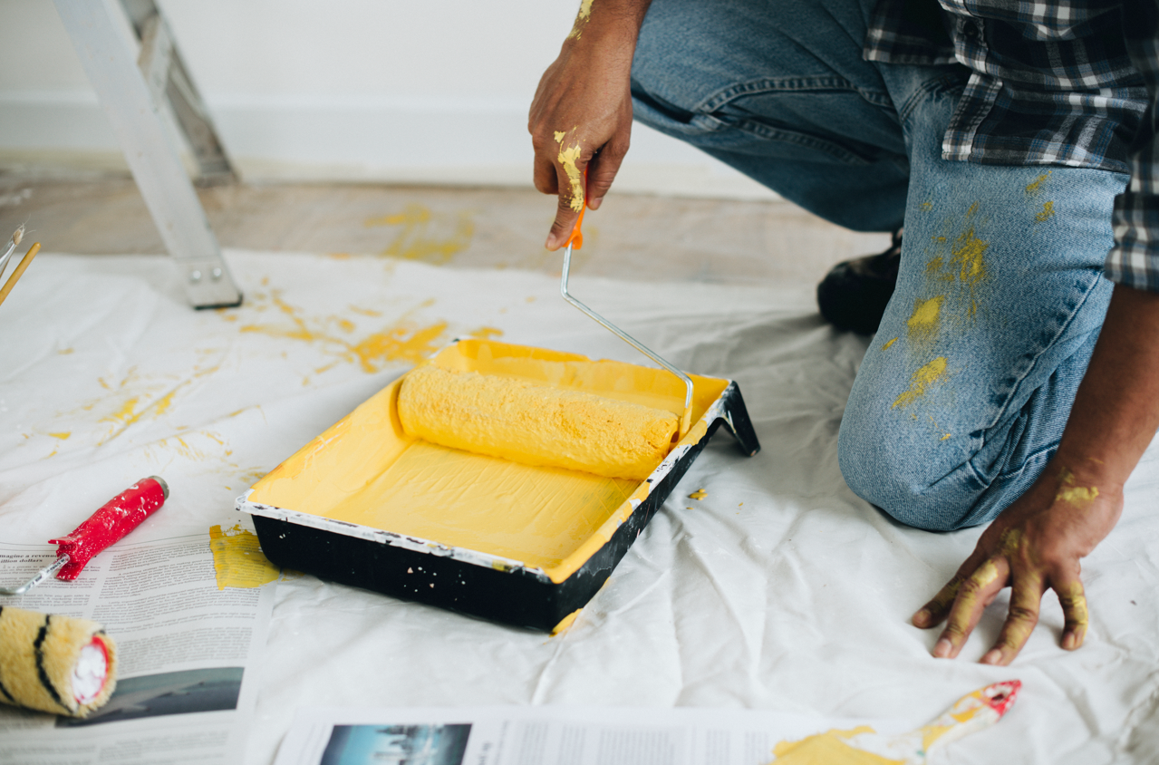 Person kneeling, painting with yellow paint roller in tray; paint splatters on jeans and newspaper.