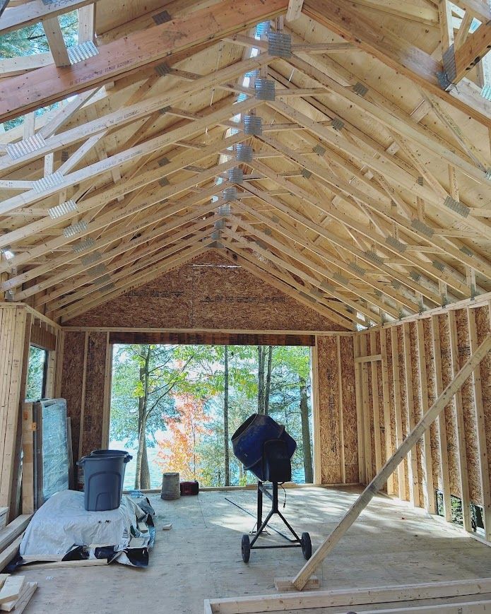 Interior view of a building under construction, showing wooden framing, roof trusses, and an open doorway with a view of trees and water.