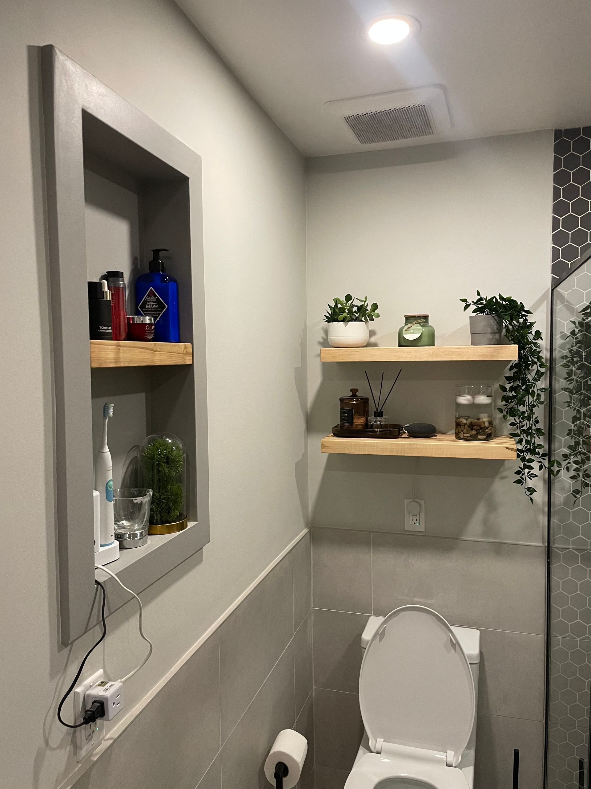 Bathroom with recessed shelf and floating shelves, holding toiletries, plants, and decor. Gray walls, white toilet.