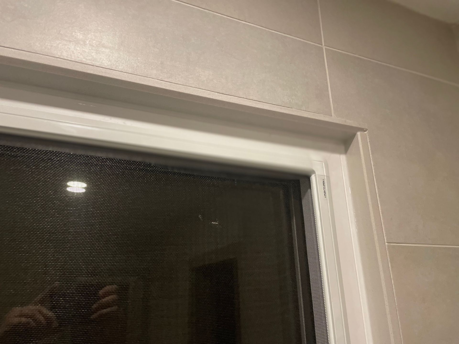 Close-up of a window with a screen and white trim against tiled wall.