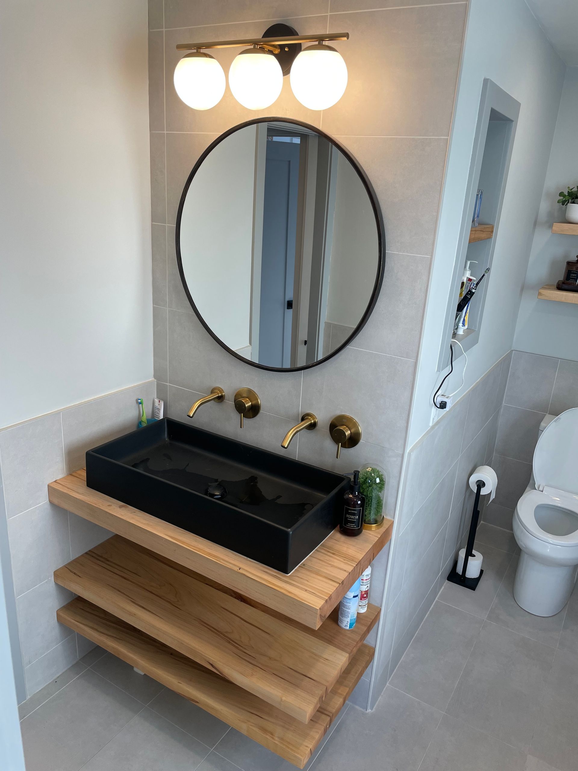 Modern bathroom with black sink, round mirror, wood shelves, and a toilet.