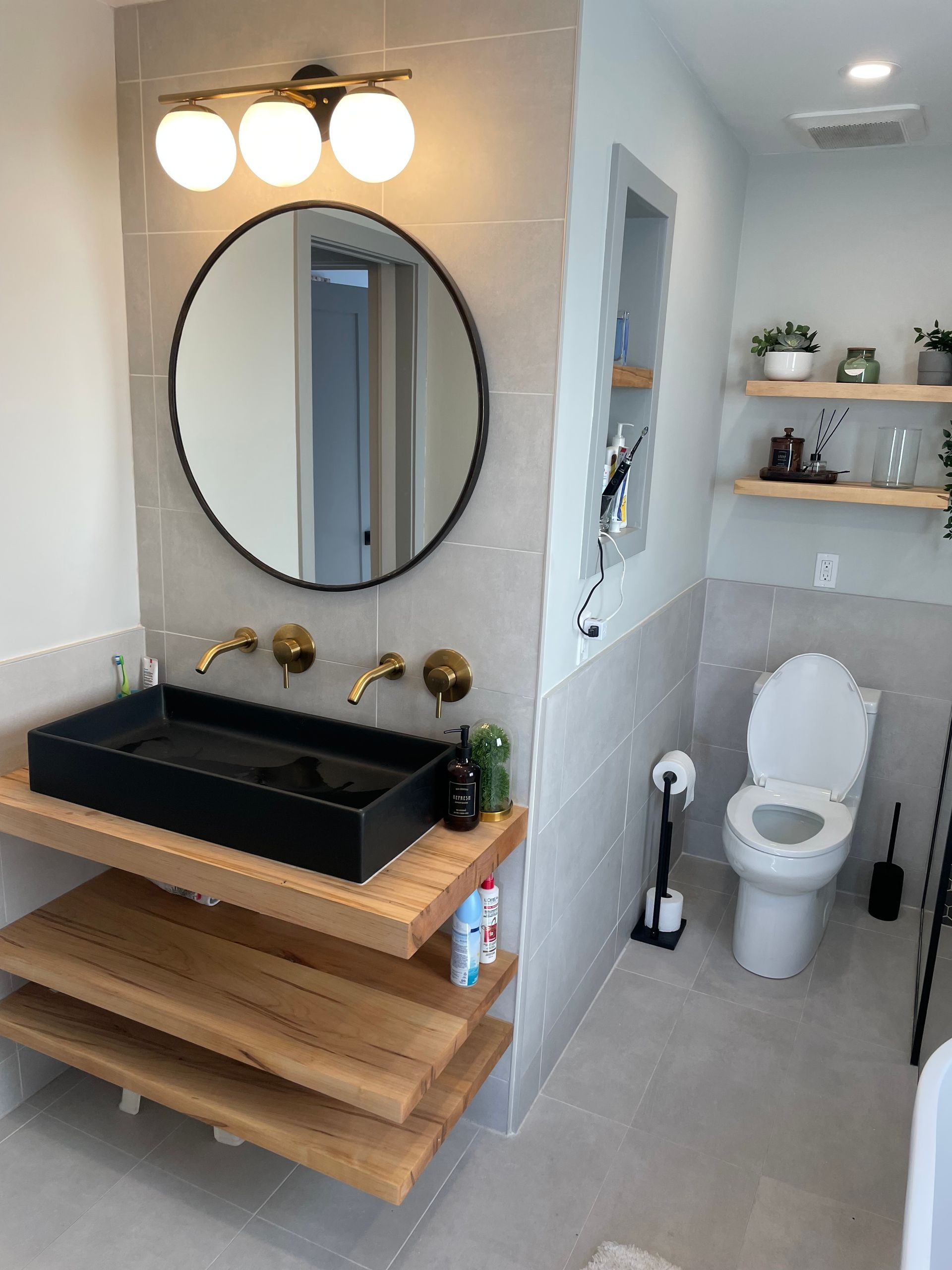 Modern bathroom with black rectangular sink on wooden shelves, round mirror, and toilet. Light gray walls.